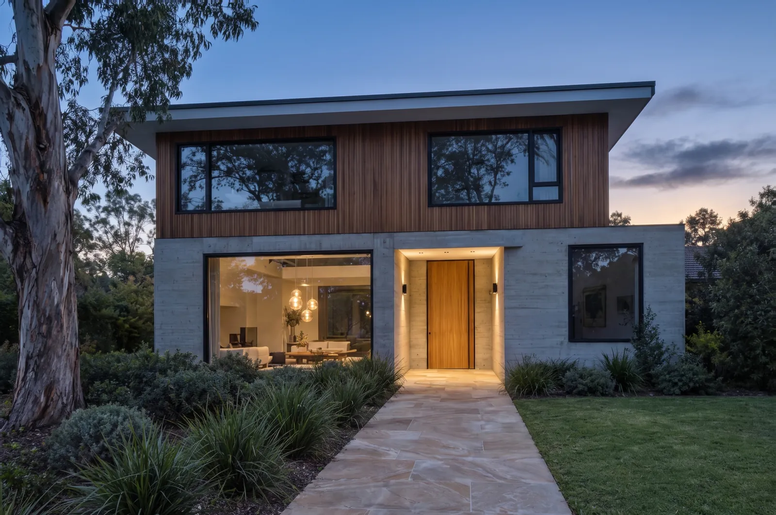 The same house edited to dusk. Only the lights already in the photo are on: two downlights over the entry, two wall lights by the front door and three pendant lights inside the large window.