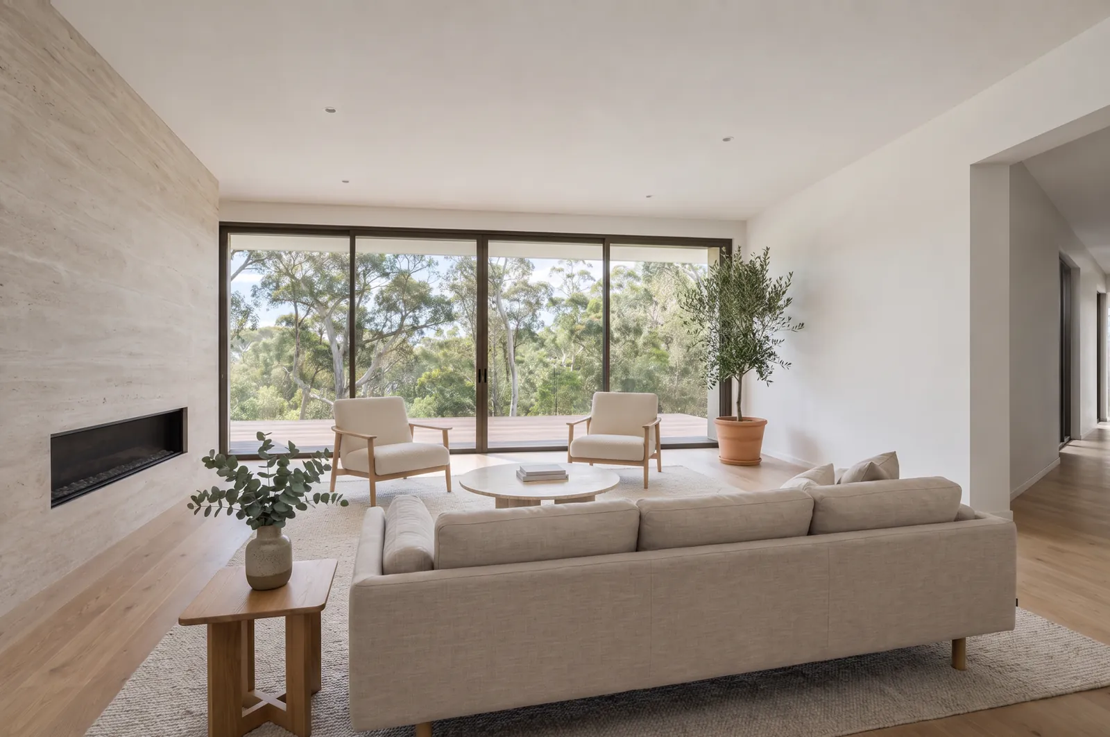 The same living room virtually staged with a linen sofa, two oak lounge chairs, a travertine coffee table, a wool rug, a side table and a potted olive tree. Walls, floor, doors and windows are unchanged.